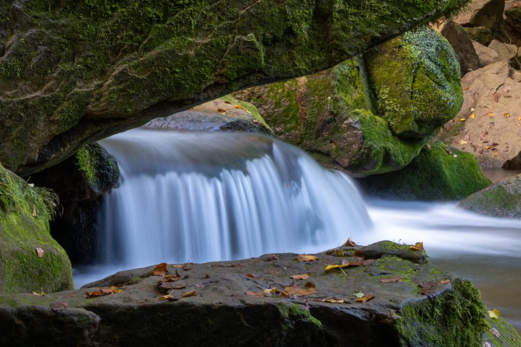 Langzeitbelichtung eines Wasserfalls unter einem moosbewachsenen Felsbogen im Müllerthal
